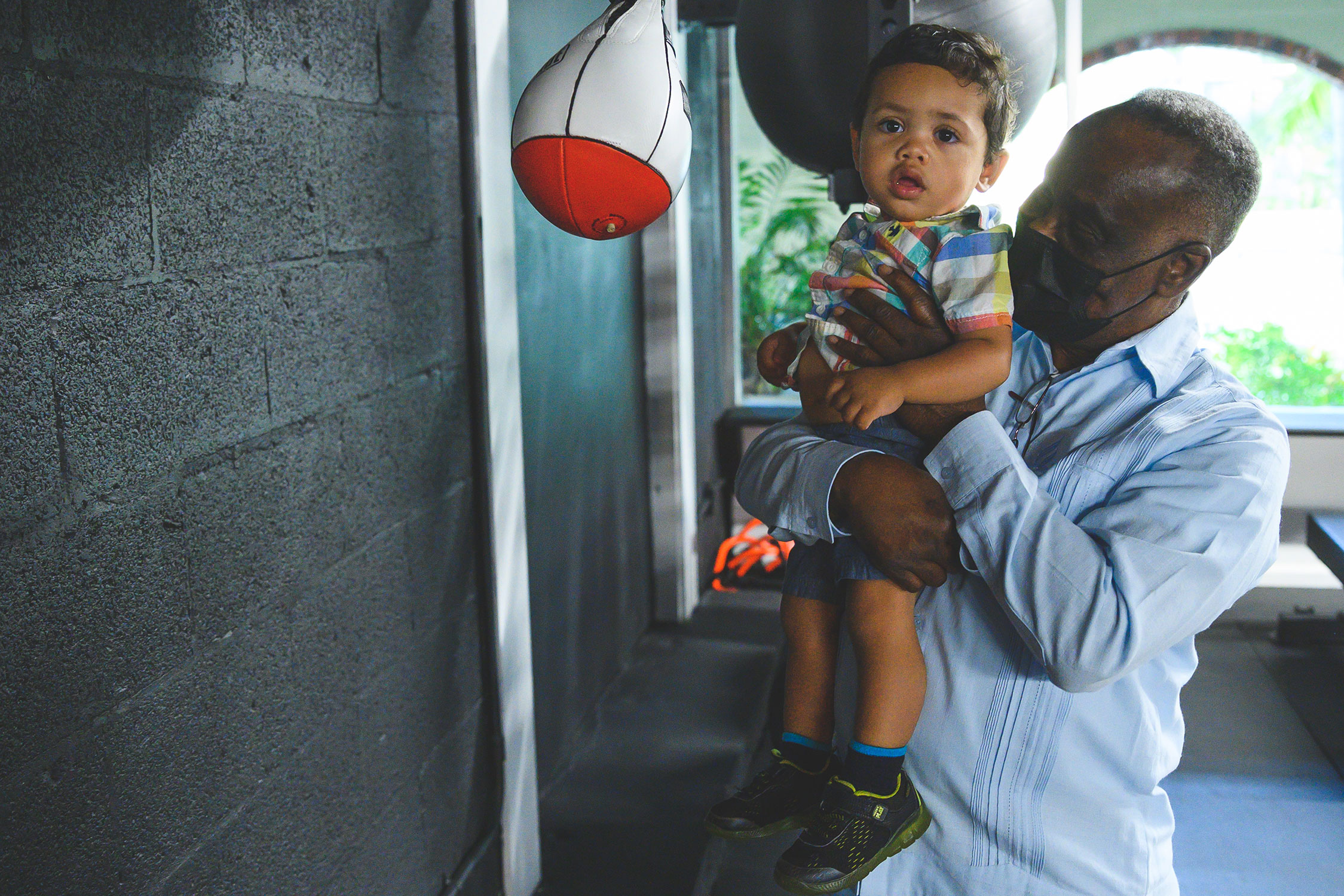 Older Black man wearing face mask holding baby up to punching bag in a boxing gym